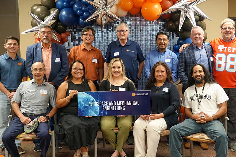 Group of attendees pose together during a UTEP Engineering homecoming event while holding a large “Aerospace and Mechanical Engineering” banner, seated and standing in rows in front of a decorative balloon backdrop and event display wall.